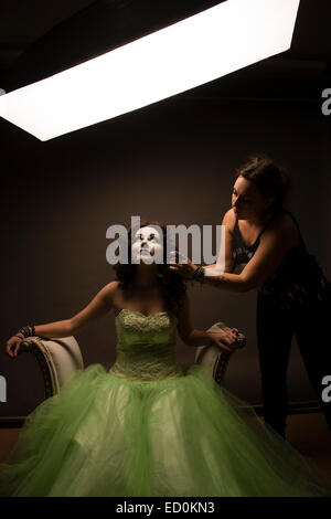 Fantasy makeover photography: A young woman girl model being made up by a make up artist to look like a porcelain white-faced painted puppet and posing like a doll in a photo studio wearing a big green frock dress ball gown Stock Photo