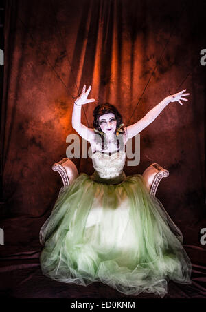 Fantasy makeover photography: A young woman girl model made up to look like a porcelain white-faced painted puppet and posing like a doll in a photo studio wearing a big green frock dress ball gown