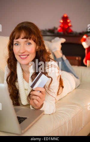 Smiling redhead lying on couch shopping online with laptop Stock Photo
