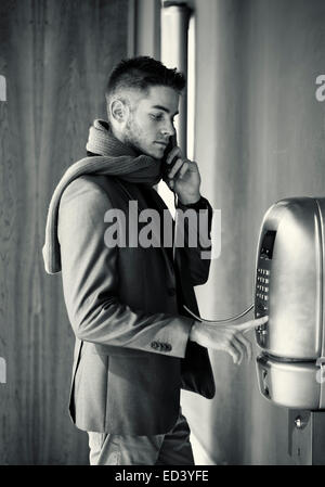 Handsome stylish young man in a tailored jacket and scarf standing using a pay phone waiting for a connection, profile view in g Stock Photo