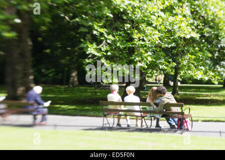 People sitting on park benches in a city; A young couple kissing and some elderly people. Stock Photo