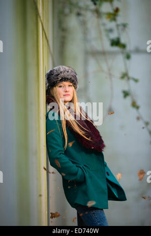 a young cool 'scandinavian' blonde woman teenage girl wearing a green coat and fur hat alone leaning back against a concrete wall outdoors  on a cold winter day UK Stock Photo