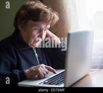 Old woman works (learning to work) on laptop in her house. Stock Photo
