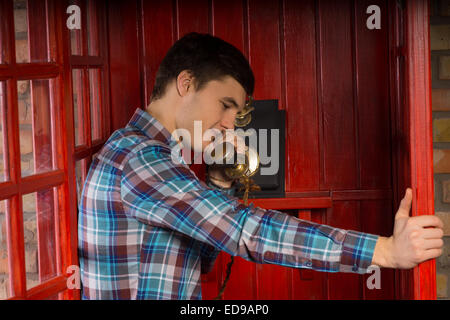 Handsome young man standing talking on a vintage telephone handset in a booth leaning on the open wooden door Stock Photo