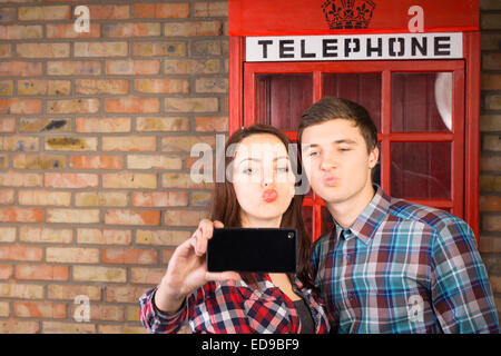 Young couple posing in front of an iconic red British phone booth taking a selfie of themselves on their mobile phone while pull Stock Photo