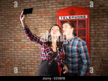 Attractive young couple taking a selfie on their mobile phone posing with an iconic red British phone booth behind them against Stock Photo