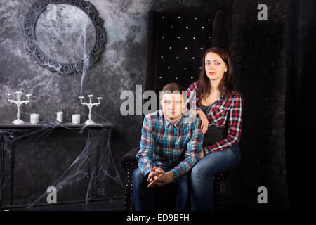 Young Couple Wearing Plaid Shirts Sitting Together in High Back Chair in Eerie Haunted House Setting Stock Photo
