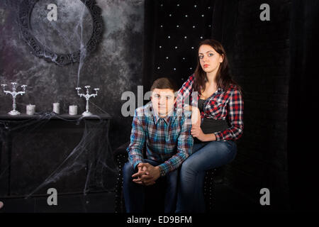 Young Couple Wearing Plaid Shirts Sitting in Black High Back Chair in Haunted House Setting Stock Photo