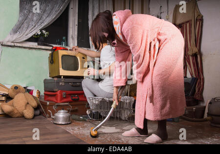 Woman in Pink Bathrobe Brushing the Floor of a Messy Abandoned Room While Partner is Busy Cleaning at the Back. Stock Photo