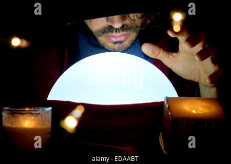 Very low key portrait of hooded man with eyes covered reading fortune on bright crystall ball, surrounded by candle light. Stock Photo