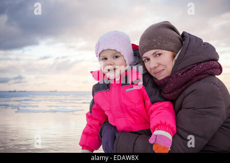 Caucasian family outdoor portrait on winter sea coast, young mother hugs her baby girl in pink jacket Stock Photo