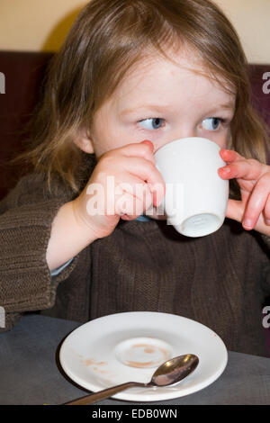 Young girl aged two years / 2 year s drinking from small coffee cup (and saucer) of babyccino baby cinno in a cafe / restaurant. Stock Photo