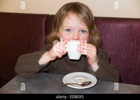 Young girl aged two years / 2 year s drinking from small coffee cup (and saucer) of babyccino baby cinno in a cafe / restaurant. Stock Photo