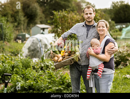 Young family standing in their allotment, smiling. Man holding a box full of freshly picked vegetables.
