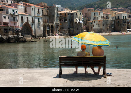 Two people sitting on a bench under a parasol overlooking the old port and ancient buildings at Cefalu Sicily Italy Stock Photo