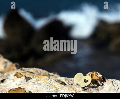 A bright beautiful metal heart is placed on top of a rock. In the background is a rocky beach and a breaking wave. Stock Photo