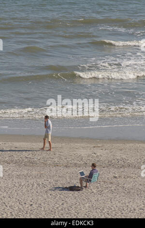 One young man works on his laptop computer while sitting on the beach as another man walks past talking on his cell phone Stock Photo
