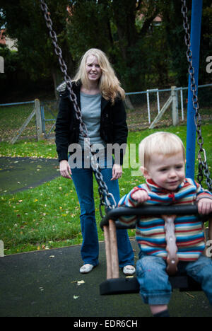 young mother pushing toddler on swing in playground Stock Photo