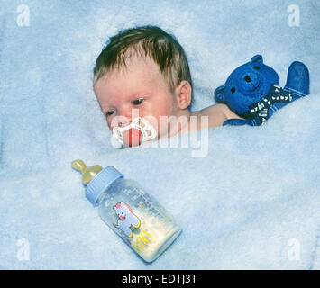 A three week old baby girl lying in a blue blanket with her teddy bear and bottle of formula. Stock Photo