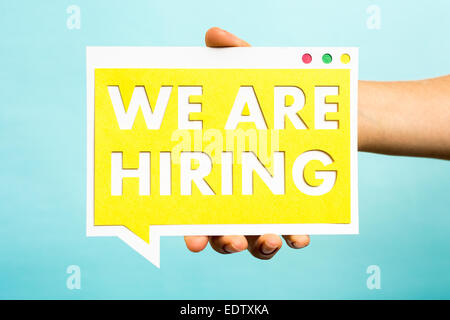 Hand holding we are hiring text with yellow paper on speech bubble and blue background. Business concept. Stock Photo