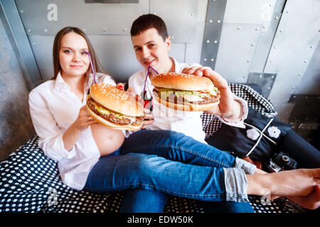 Young happy pregnant family resting and eating fast food. Fresh cheeseburger and soda water. Wide angle shot, selective focus. Stock Photo