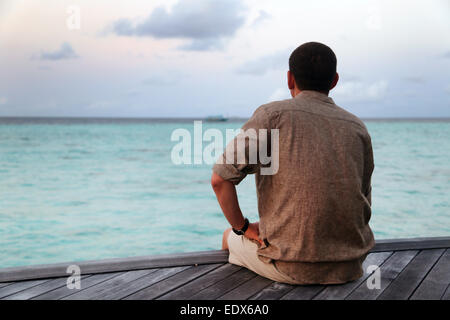 A lonely man is sitting on a wooden pier and looking into the ocean distance. Stock Photo