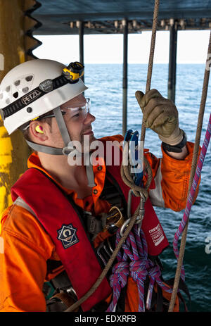 Close-up portrait of an IRATA rope access technician in white helmet and orange coveralls, with rigging hardware, on an offshore structure. Stock Photo