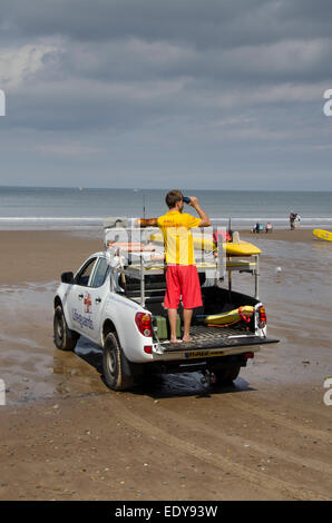 Young RNLI lifeguard man on duty watching people with binoculars in sea, stands on back of pick-up truck - Whitby beach, North Yorkshire, England, UK. Stock Photo