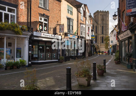 Looking down narrow, historic Kirkgate, lined with small shops, towards a tower on the west front of Ripon Cathedral - North Yorkshire, England, UK. Stock Photo