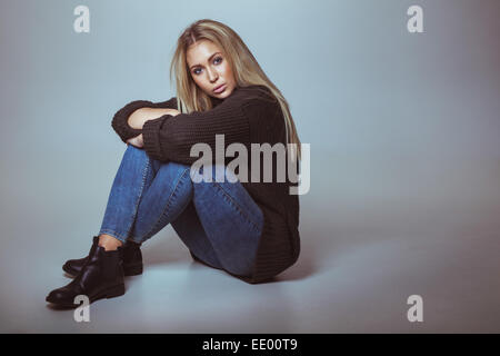 Image of attractive woman wearing sweater sitting on floor looking at camera. Blond woman in studio with copy space. Stock Photo