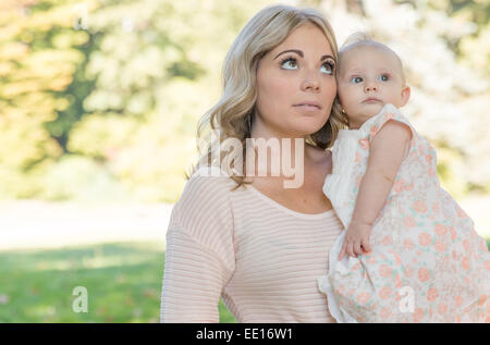Young blond mother holding six month old baby girl looking up in the sunlit park Stock Photo