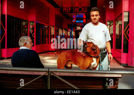 Man with a muzzled Boxer dog in front of a Peep Show in Madrid, Spain, next to a senior man on a bench Stock Photo