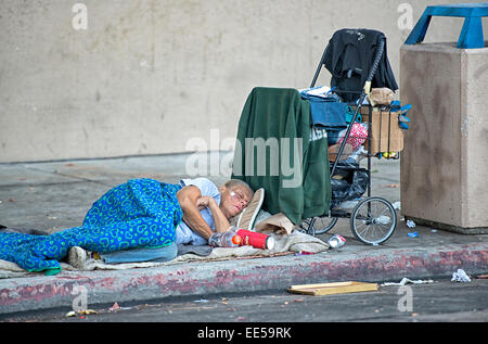Homeless Elderly Woman Sleeping on Sidewalk Next to Shopping Cart and Trash Container, East Village, San Diego, California USA Stock Photo
