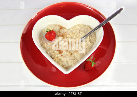 Porridge/oatmeal with honey cinnamon in a heart shaped bowl on a red plate with a strawberry garnish. On white wood background. Stock Photo