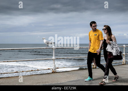 A young couple walk along a British seaside pier, holding hands on a cloudy day. Stock Photo