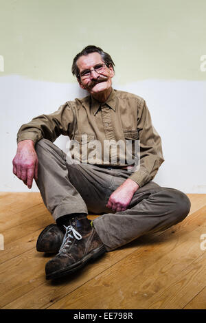 Man in his 60s with moustache wearing khaki shirt and trousers sitting on the floor. Stock Photo