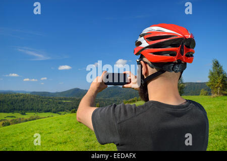 Cyclist taking pictures with smart phone. Caucasian man in bike helmet taking smartphone photo of mountains. Outdoor activity. Stock Photo