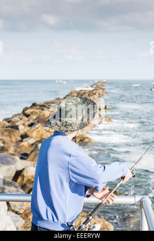 Man with sun hat seen from side standing at a railing with fishing rod in hand looking out at the ocean, stone jetty and boats. Stock Photo