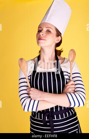 Portrait of an attractive female chef over a yellow background holding two wooden spoons. Stock Photo