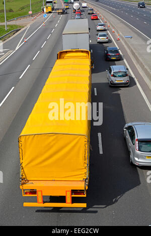Aerial back top view yellow hgv articulated lorry truck & trailer slow moving motorway traffic jam using two lanes obstructions ahead Essex England UK Stock Photo