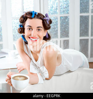 Young woman with hair curlers drinking coffee sitting on the couch at white domestic background Stock Photo
