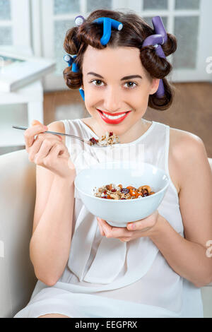 Young woman with hair curlers eating granola breakfast sitting on the couch at home Stock Photo