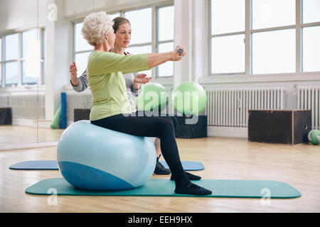 Female trainer assisting senior woman lifting weights in gym. Senior woman sitting on pilates ball doing weight exercise. Stock Photo