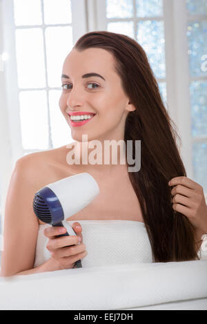 Young woman with long and dark hair drying hair with hairdryer in the bathroom Stock Photo
