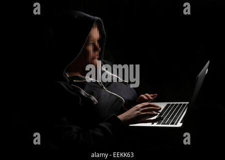 Person wearing a hoodie using a laptop in a dark room with their face pixelated depicting a hacker undertaking an illegal act Stock Photo