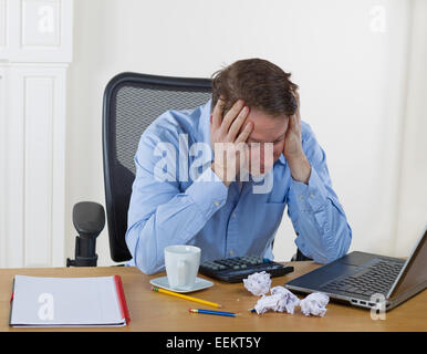Mature man holding head in his hands while looking down at desktop with laptop, calculator, broken pencils, wadded paper, coffee Stock Photo