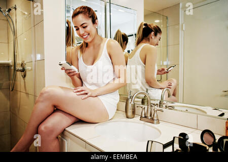 Caucasian woman using cell phone on bathroom counter Stock Photo