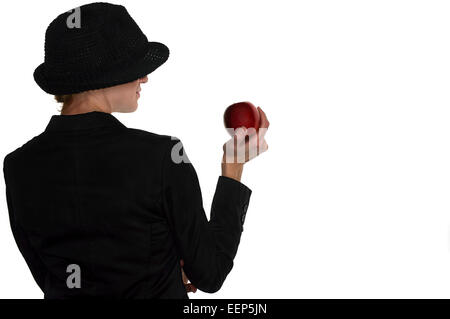 Mysterious woman wear all black hides her face with a black hat faces backward holding up a red apple at her side. Stock Photo