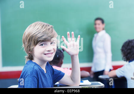 Happy little schoolboy raising hand while sitting at desk with classmates studying in background. Stock Photo