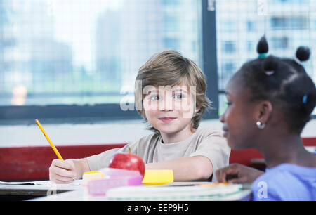 Cute caucasian kid at classroom desk drawing and smiling to his afro american female friend. Stock Photo